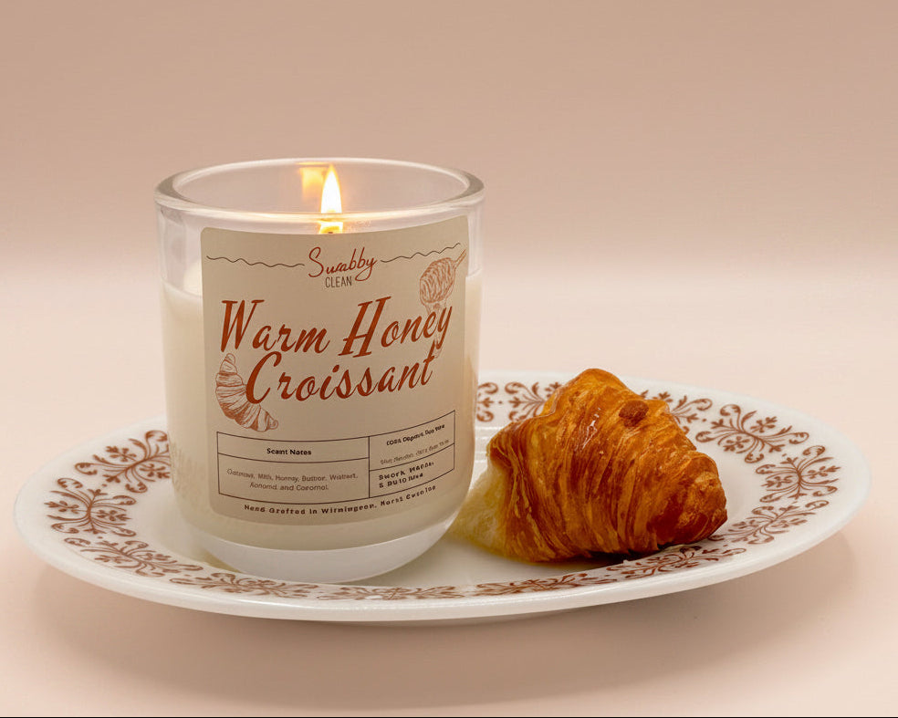 Candle labeled 'Warm Honey Croissant' on a plate with a croissant, next to a mug with 'See The Good' text.